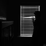 Silhouette of a person looking through window blinds in a dark room. Mystery and introspection.